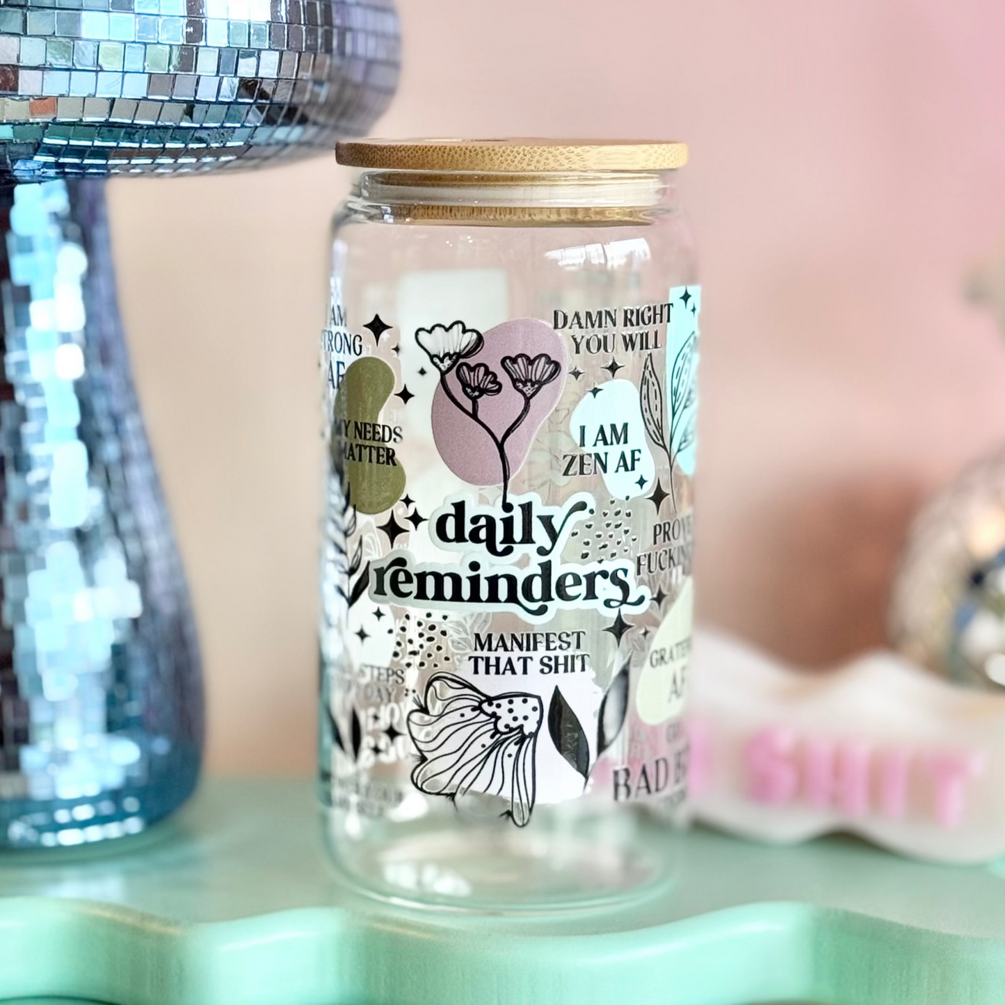 Glass jar with decorative text and illustrations on a light blue stand with a pink background