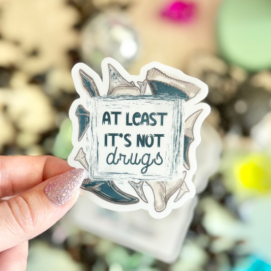 Hand holding a sticker with text 'At least it's not drugs' against a festive background with confetti and decorations.