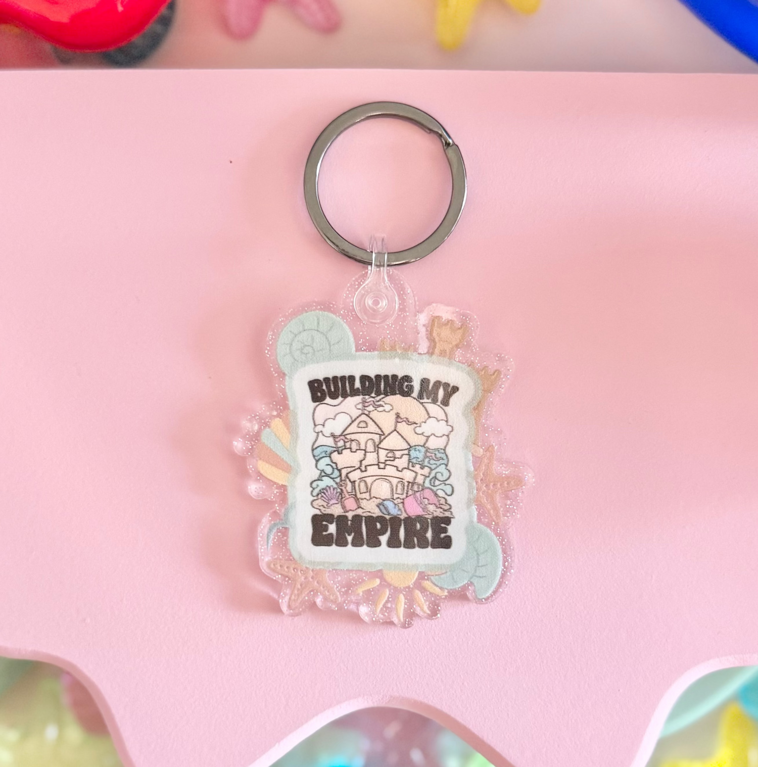 Keychain with 'Building My Empire' design on a pink card surrounded by colorful decorative items.